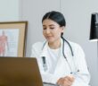 Junge Ärztin sitz vor ihrem Laptop. Um den Hals hängt ihr ein Stethoskop