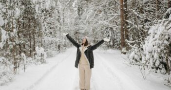 Frau steht auf winterlichem Weg im Wald und reckt die Arme in die Höhe.